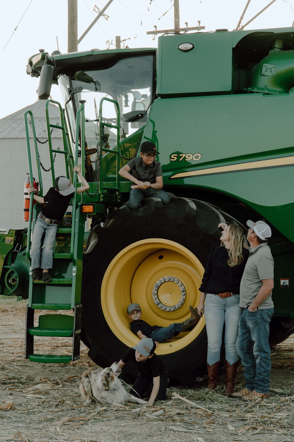 Group gathered around the massive wheel of a green combine harvester with dogs nearby.
