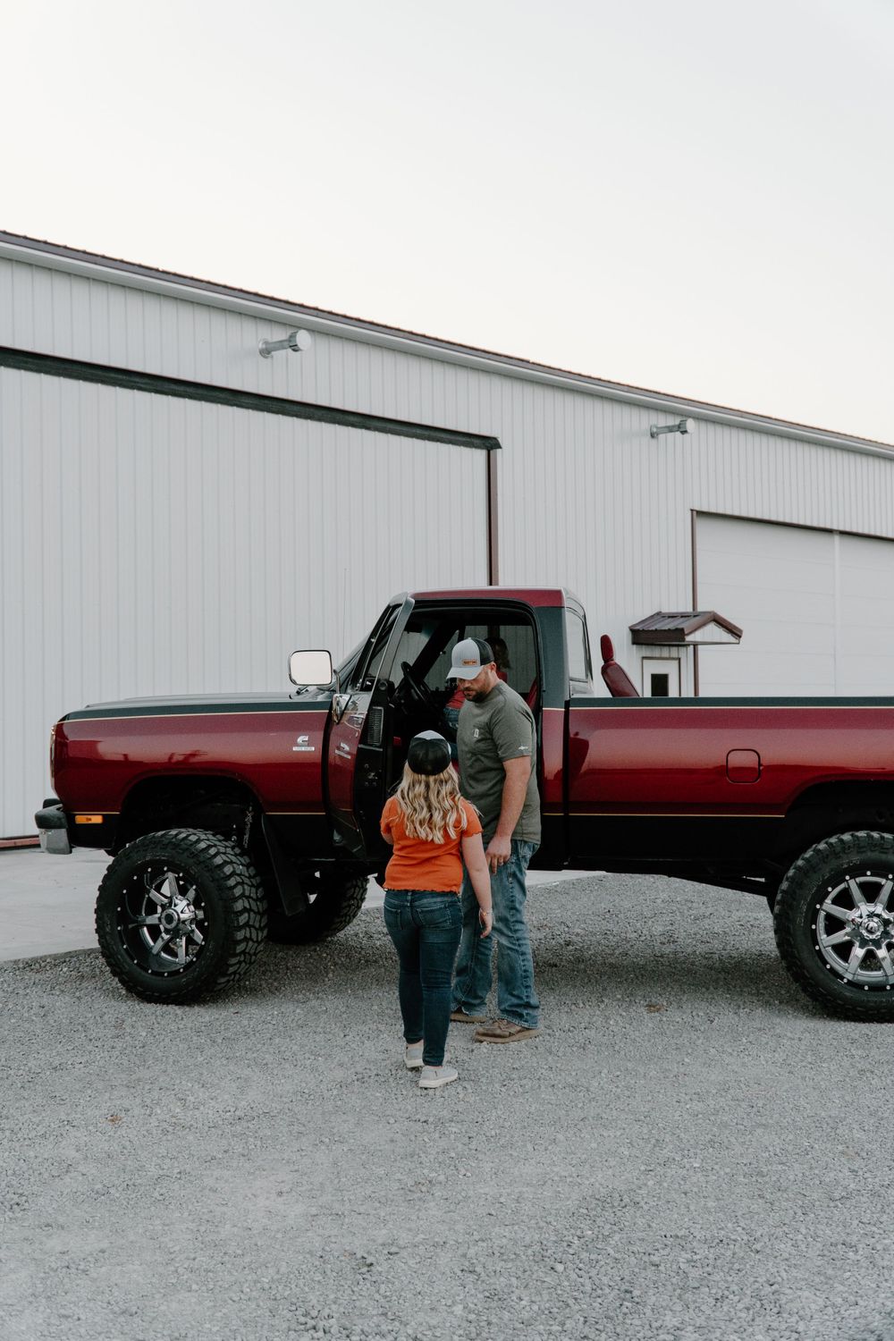 A lifted red pickup truck is showcased next to people on a gravel lot by industrial buildings.