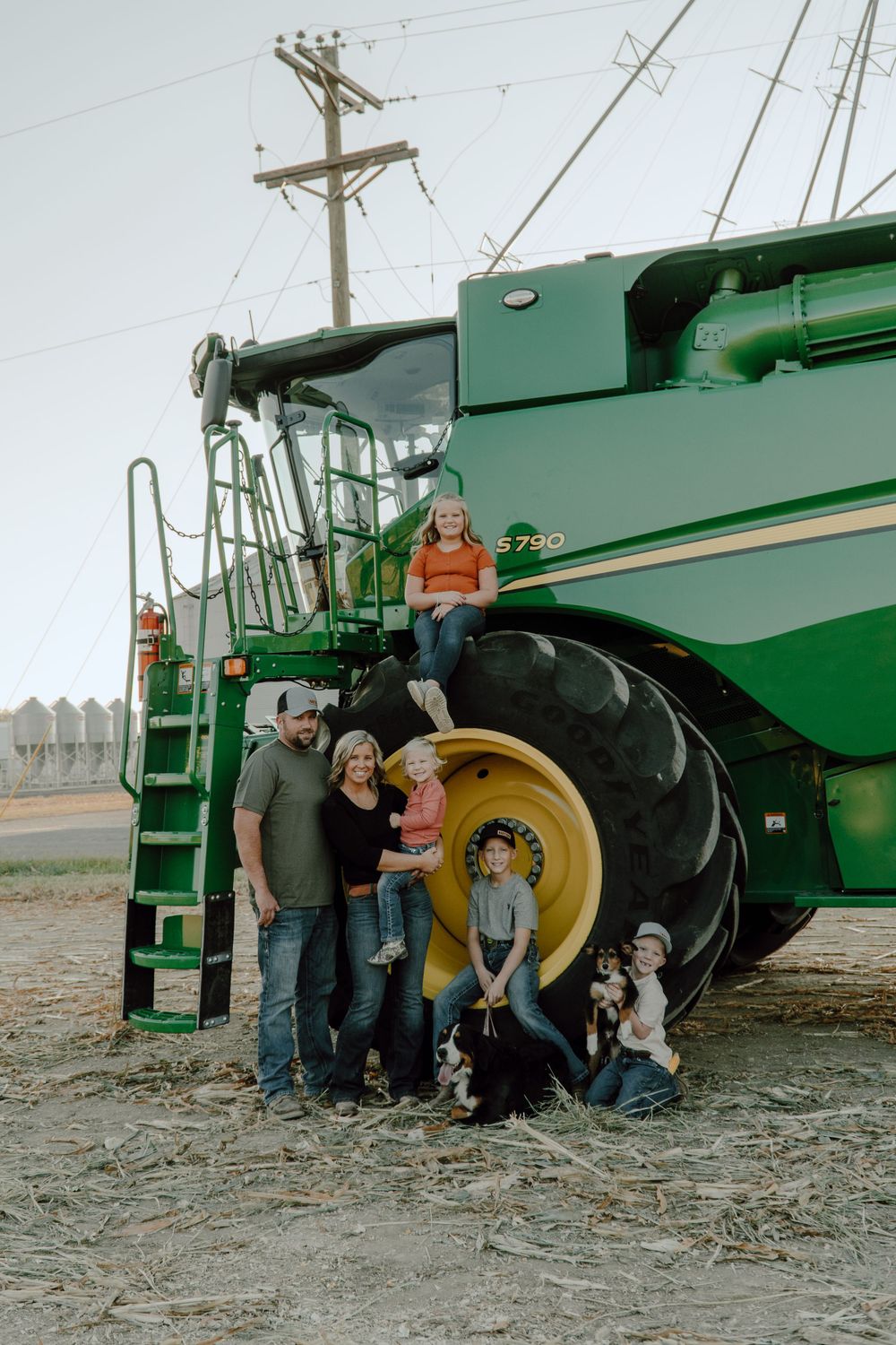 Family and dogs pose in front of a large green combine harvester with grain silos in background.