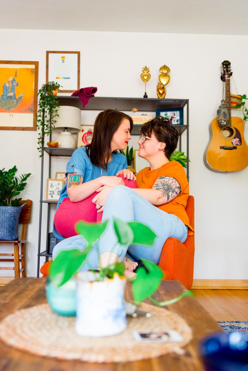 two woman in colorful clothing and colorful apartment sitting on an orange chair, kissing