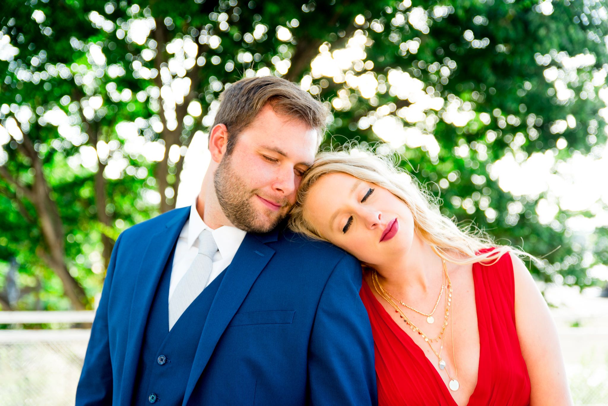 blonde woman in red dress and man in navy suit lean on each other with eyes closed in front of big tree