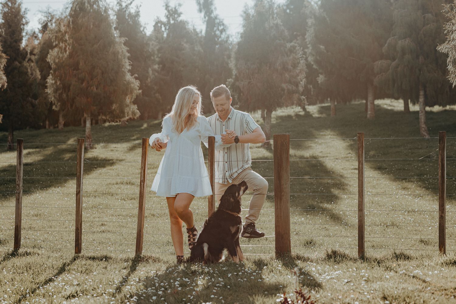 Couple leans against wooden fence posts with their loyal black dog in golden sunlight.