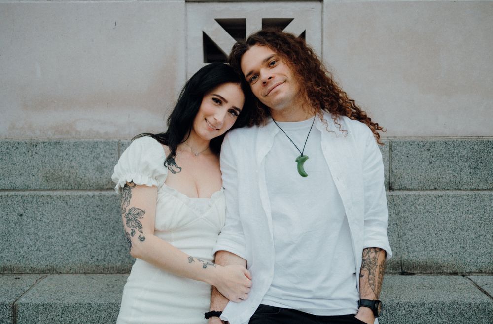 A couple in casual white outfits poses against a concrete wall with geometric art.
