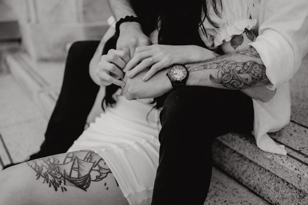 Close-up of tattooed hands with a watch resting on legs in black and white.
