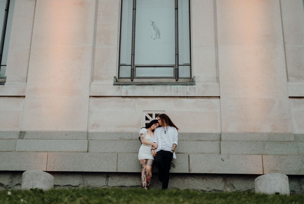 A couple shares a tender moment against a white stone building wall at dusk.
