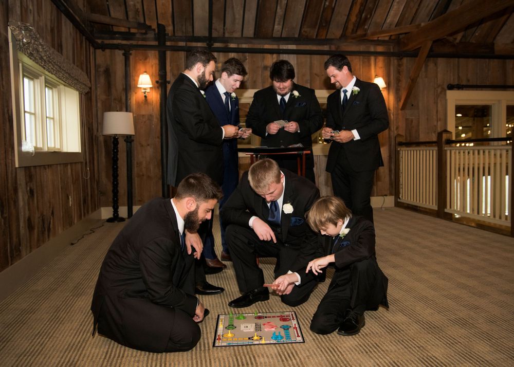 Groomsmen in black suits gathered around playing a board game in rustic wooden venue.