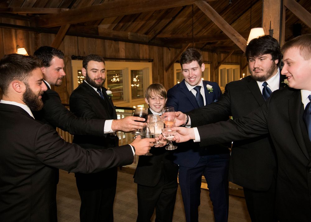 Group of groomsmen in dark suits toasting with drinks in rustic barn setting.