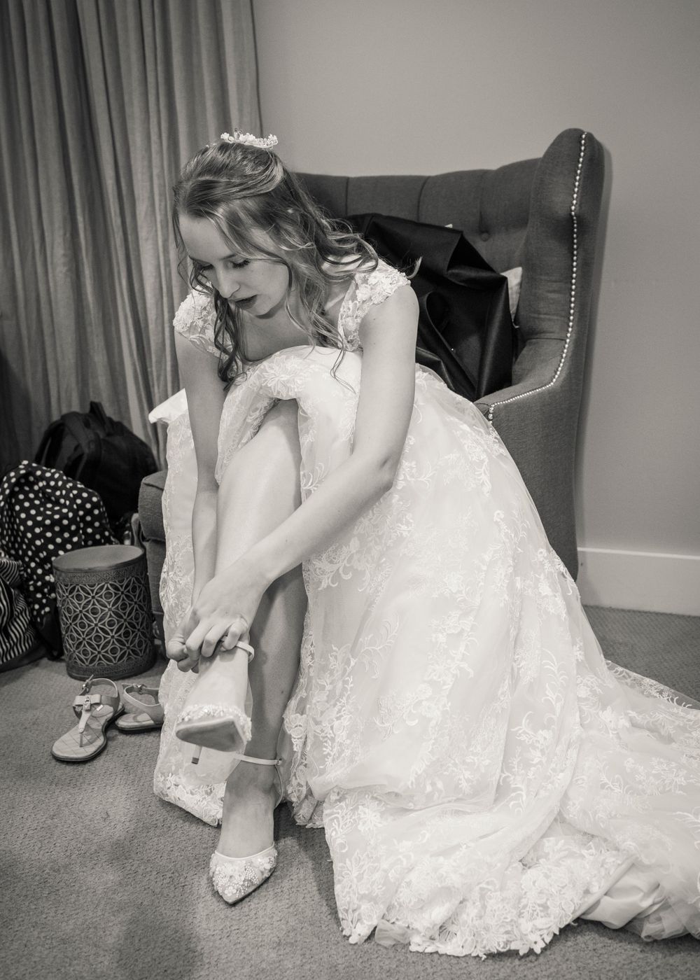 Person in white lace wedding dress putting on shoes while seated in grey armchair.
