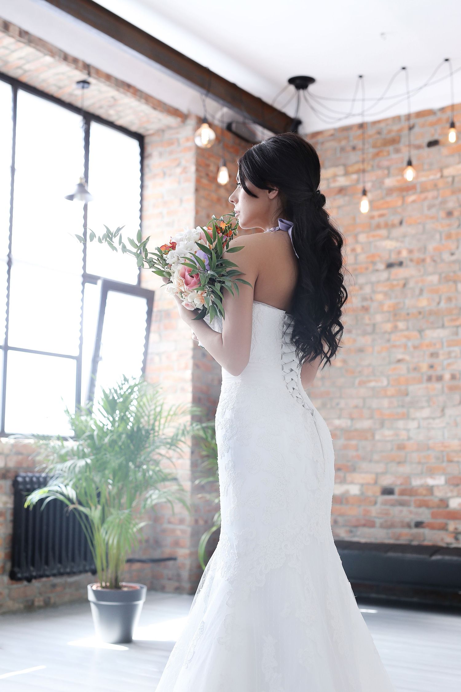Bride in fitted mermaid wedding dress with long dark hair holds bouquet in industrial loft space.