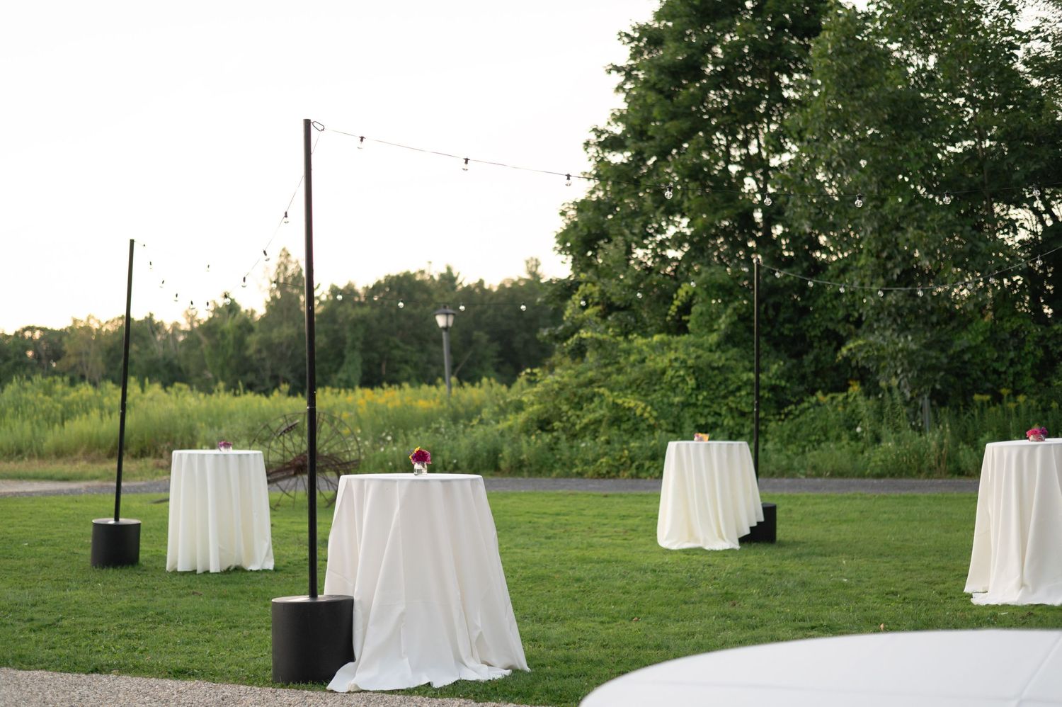 White cocktail tables with linens set up outdoors with string lights overhead.