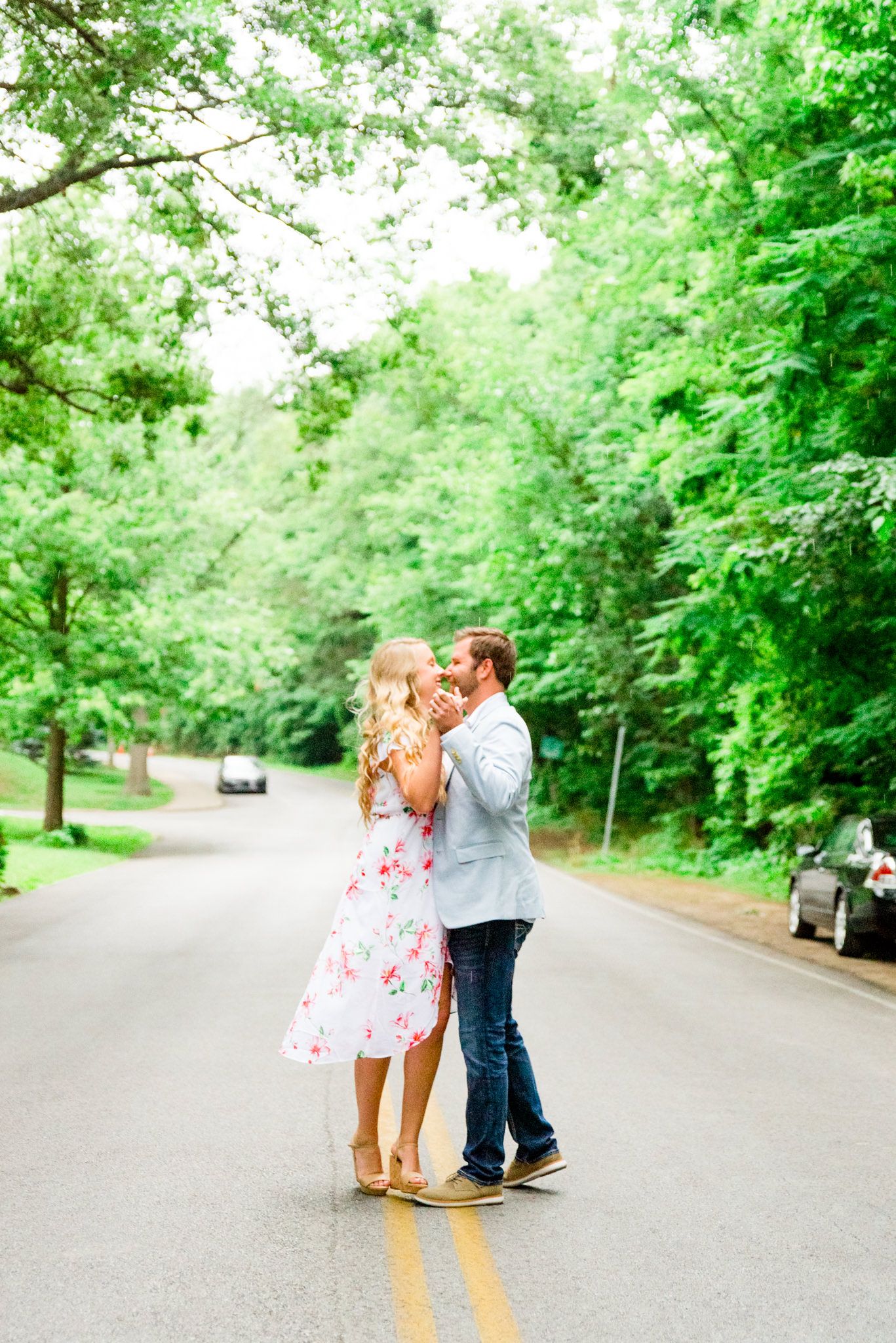 engaged couples in floral dress and light blue suit jacket and jeans dancing in middle of road under trees