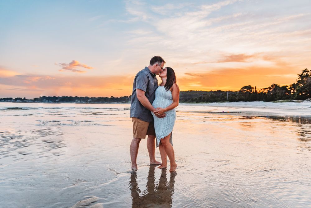 A couple stands ankle-deep in beach water during sunset for their maternity photos.