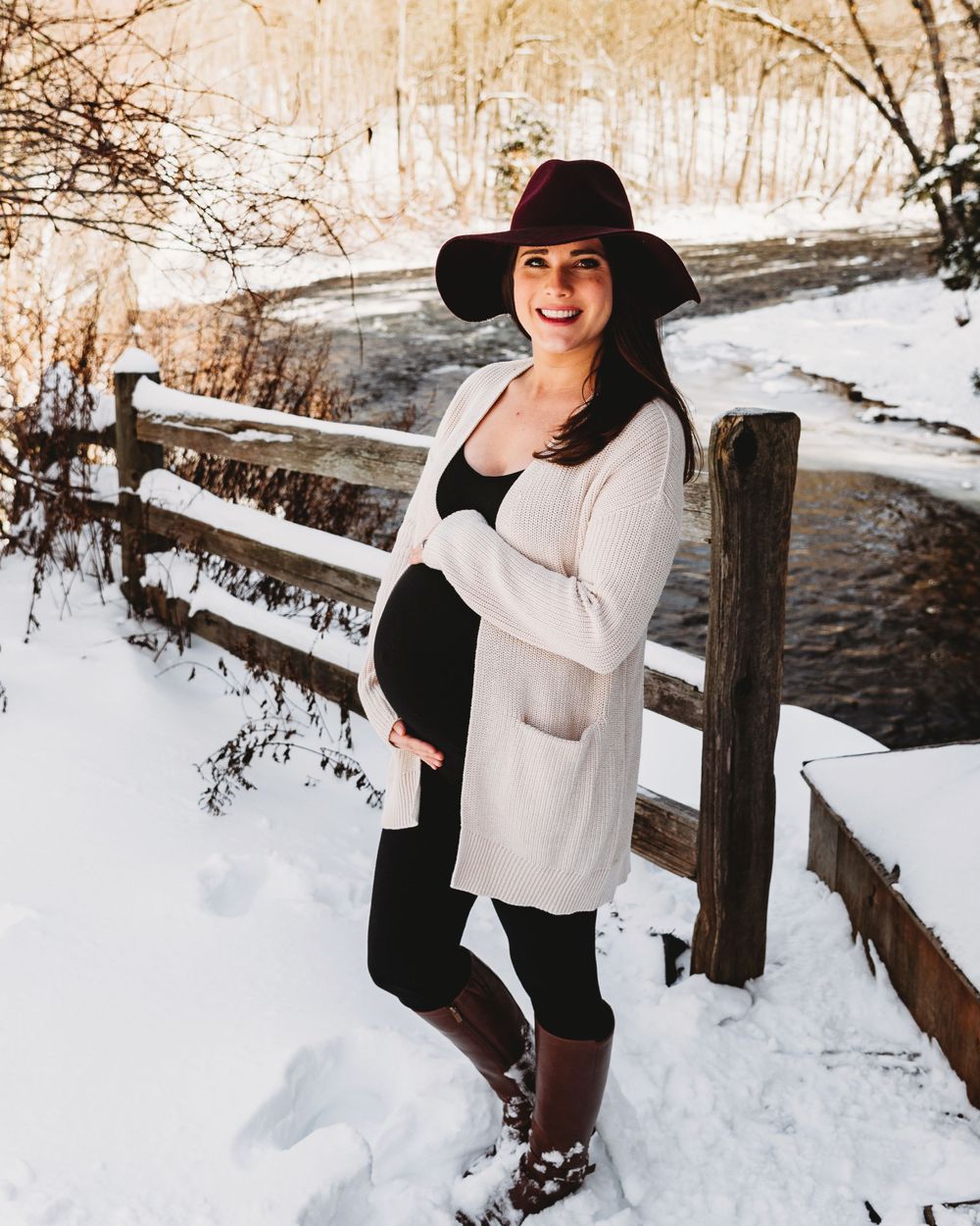 A pregnant woman in a winter hat and sweater poses by a snowy wooden fence.