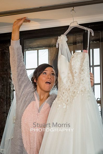 bride in a onesie stretches to reach and lift down her wedding dress hanging in a window at abbey house hotel