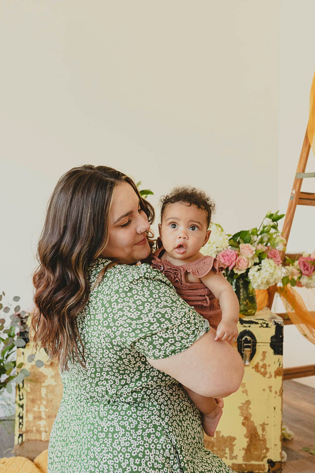 6 month old baby girl in her mom's arms. Mom is smiling into her cheek. Pink flowers surround them.