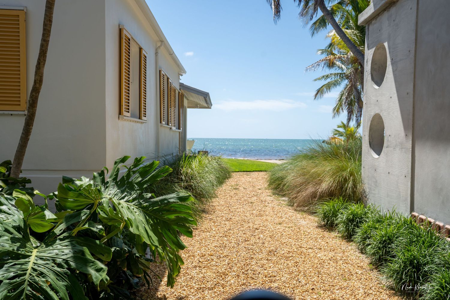 Shell-lined garden path between white buildings leads to beach and palm trees.