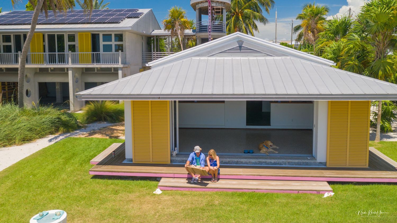 Modern yellow Florida home with metal roof and palm trees against blue sky.