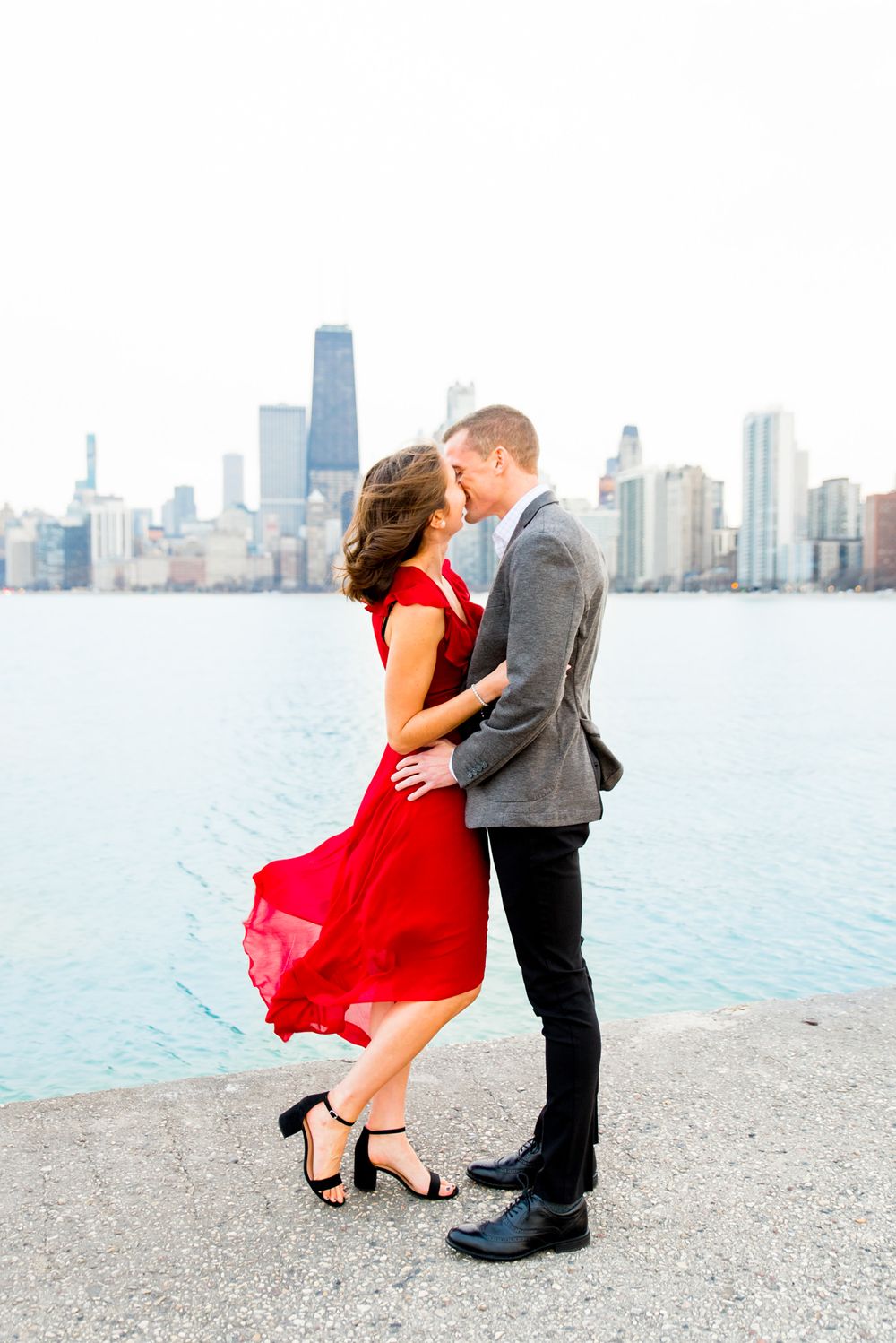 woman in flowing red dress kissing fiance for engagement pictures at North Avenue Beach on windy day