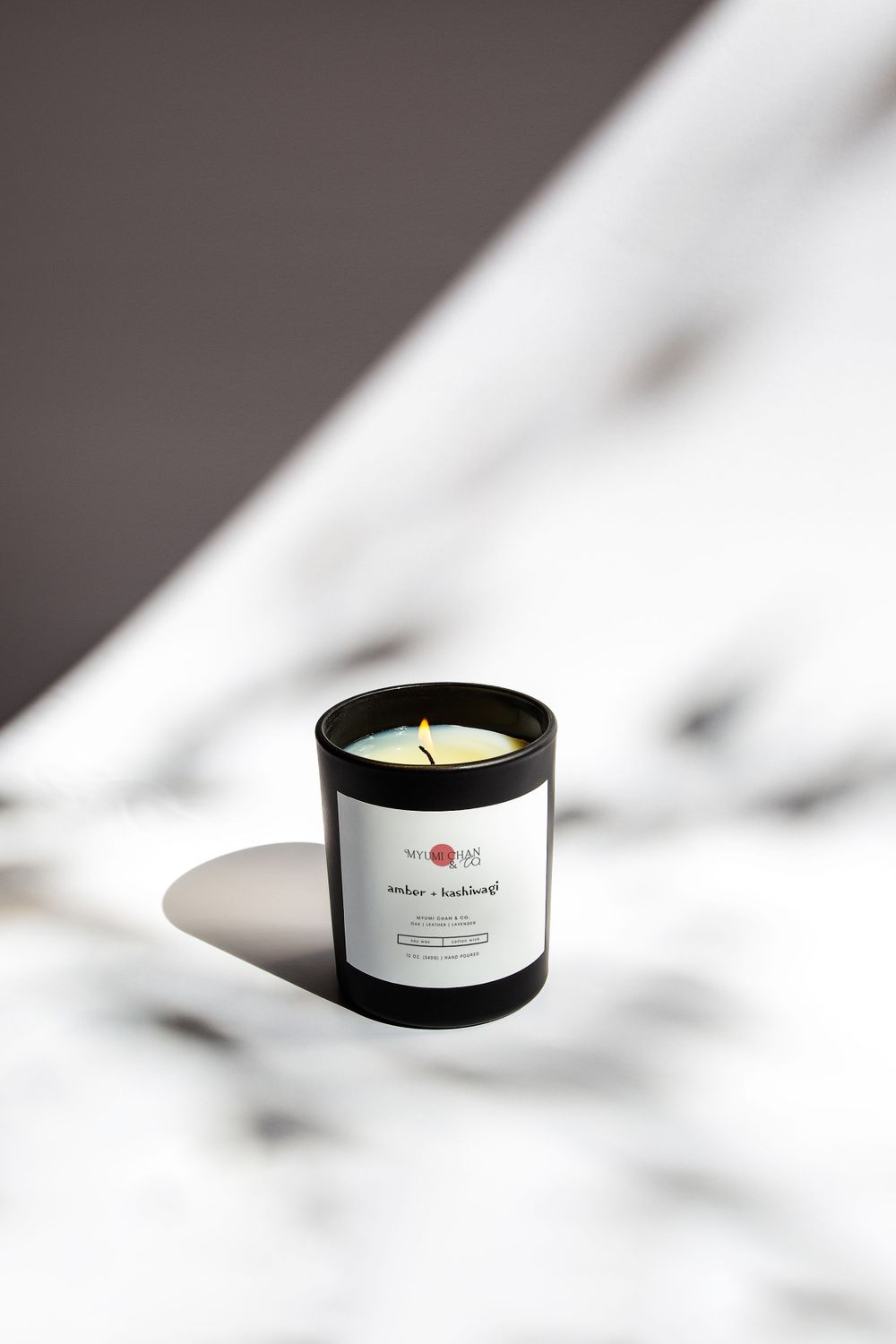 Black scented candle with white label casting dramatic shadows on white surface.