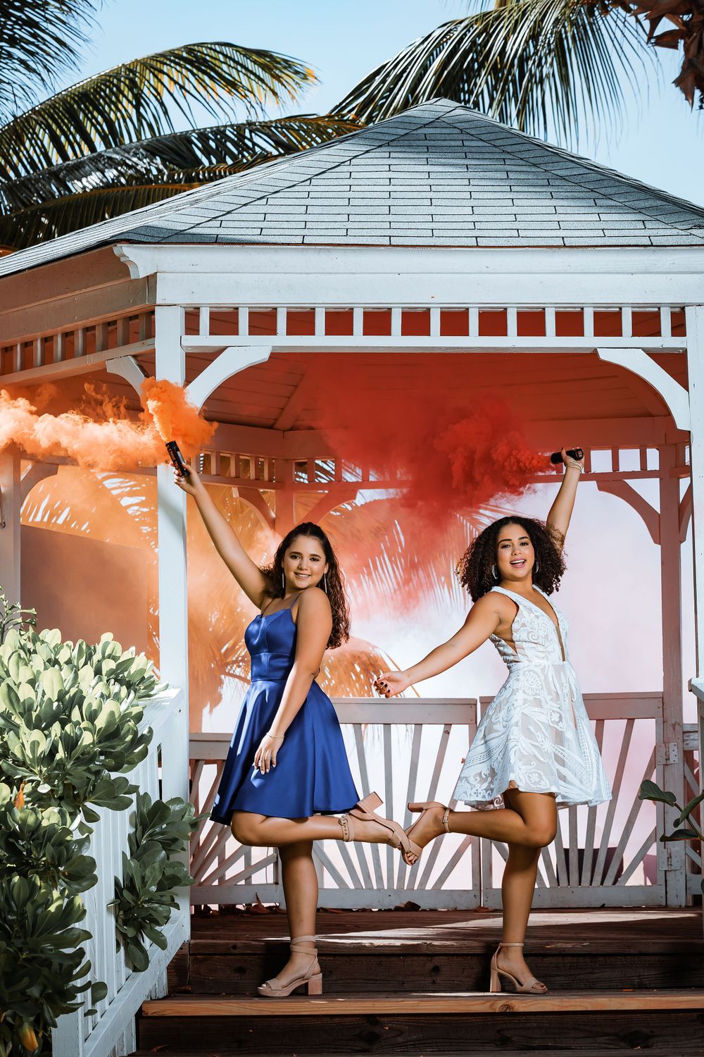 Two figures pose in a white gazebo with tropical palm trees and orange smoke effects.