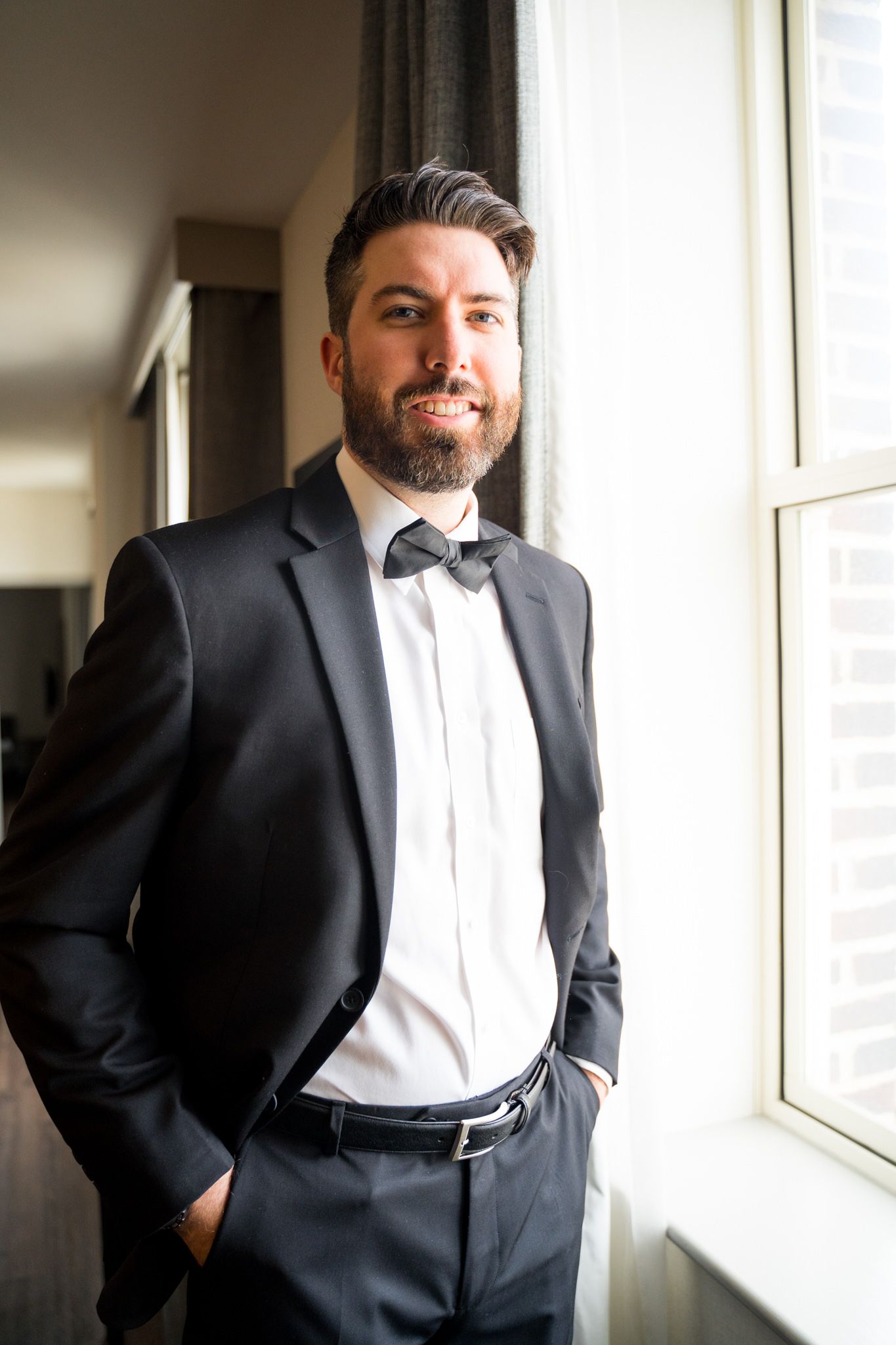 brunette groom with beard in black suit and bowtie smiling next to window in for downtown St. Louis wedding