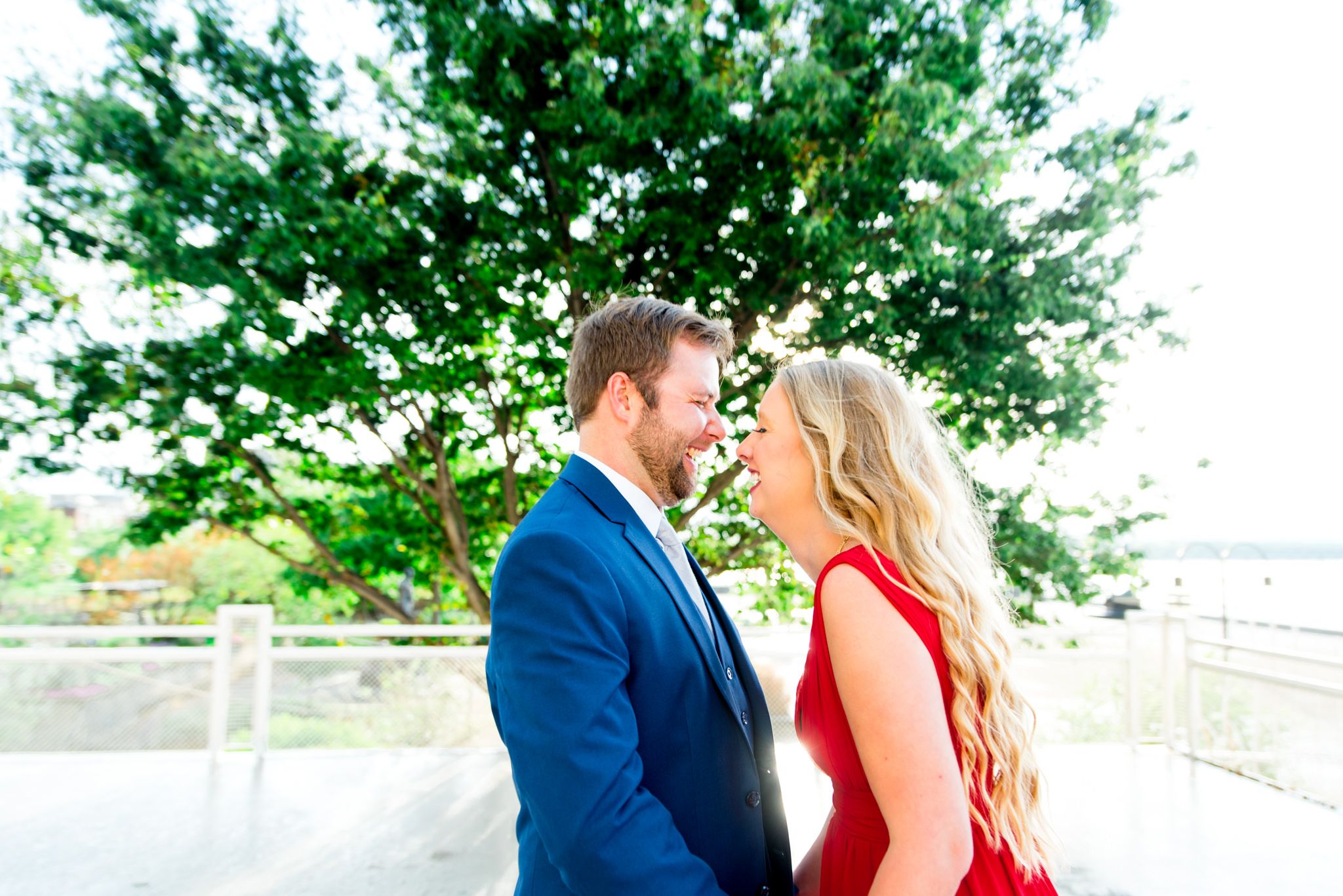 blonde woman in red dress and man in navy suit laugh with each other with eyes closed in front of big tree