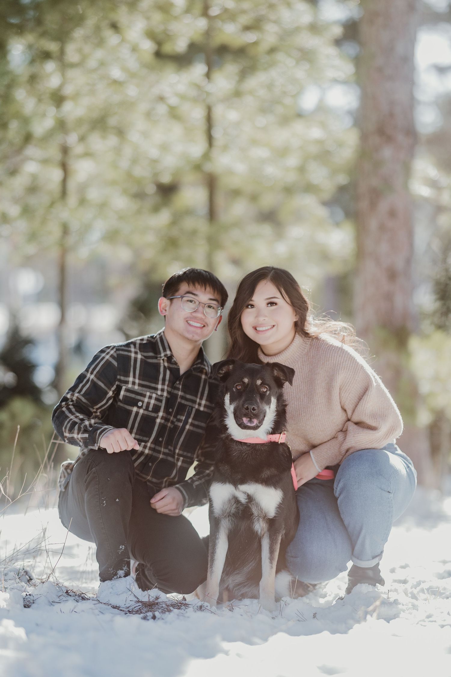 A winter portrait of a couple with their black and white dog sitting in the snow.