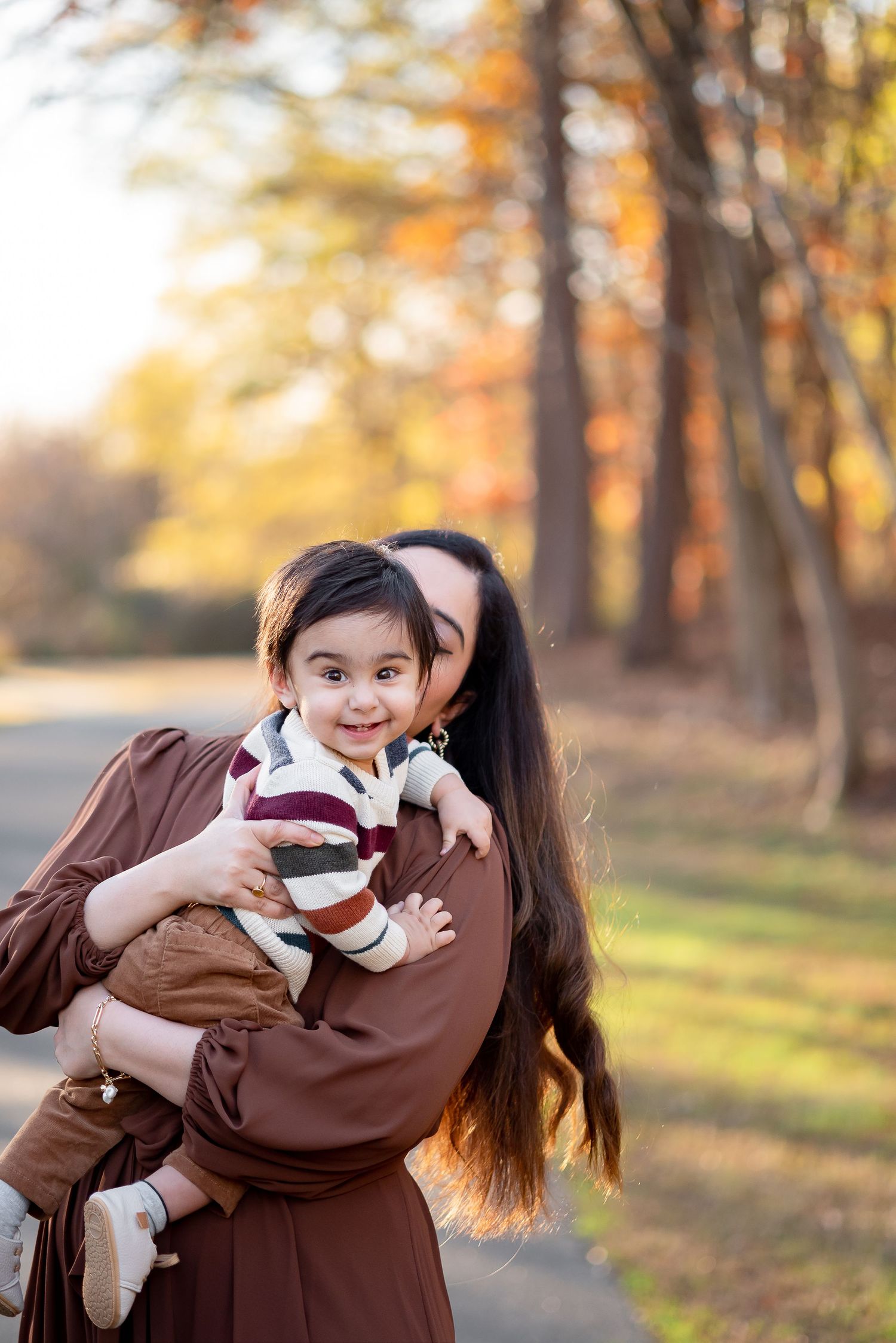 Outdoor fall portrait of a mother lifting her smiling toddler boy, surrounded by golden autumn trees and soft natural light.