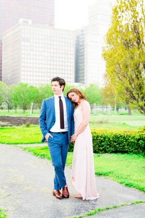 man in navy suit and woman in light pink dress stand with hands held in Grant Park Chicago for sunset engagement picture