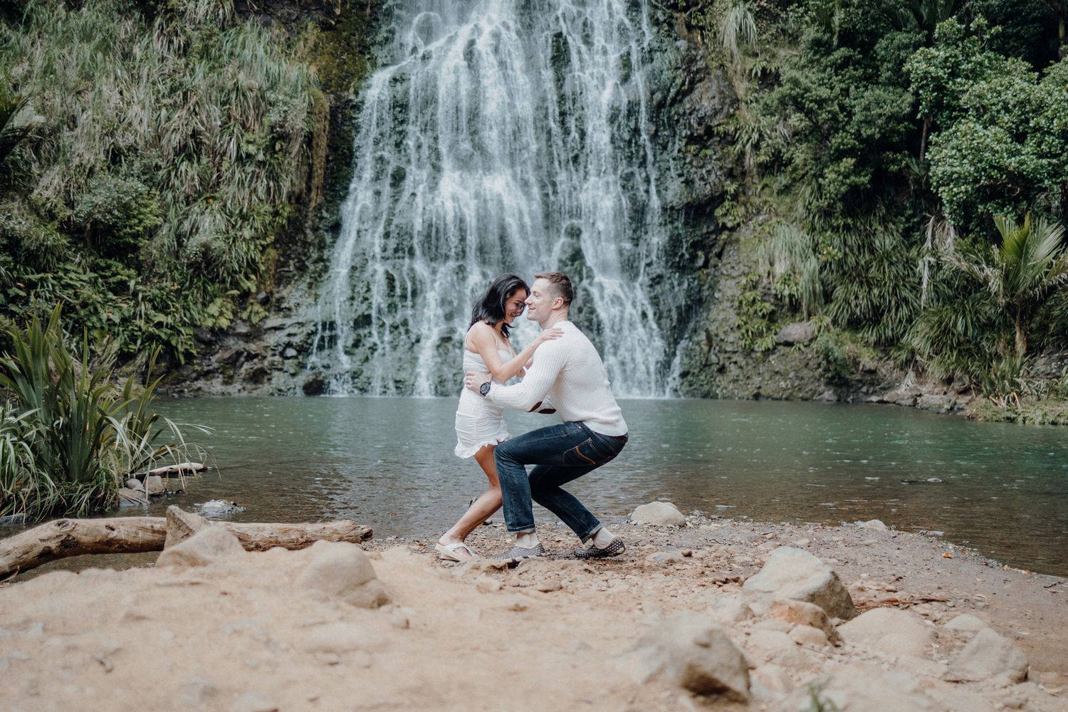 A romantic couple embraces in front of a stunning waterfall on rocky terrain.