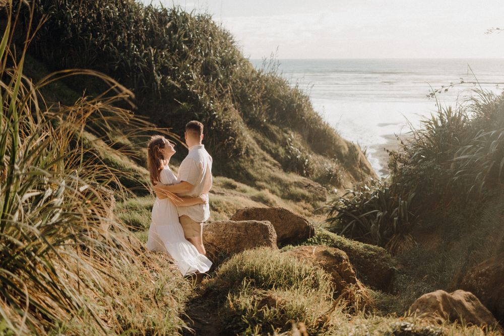 A couple in white clothing embraces on a grassy coastal path during golden hour.