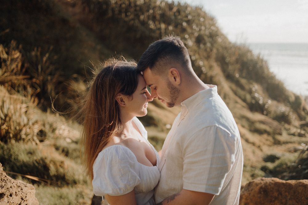 A romantic coastal photoshoot featuring a couple in white outfits sharing an intimate moment on a hillside.