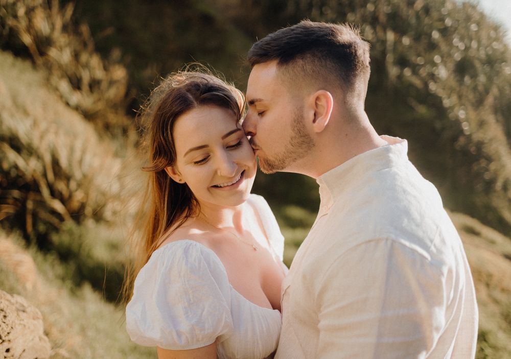 A tender moment between a couple in white clothing against a scenic coastal backdrop at sunset.