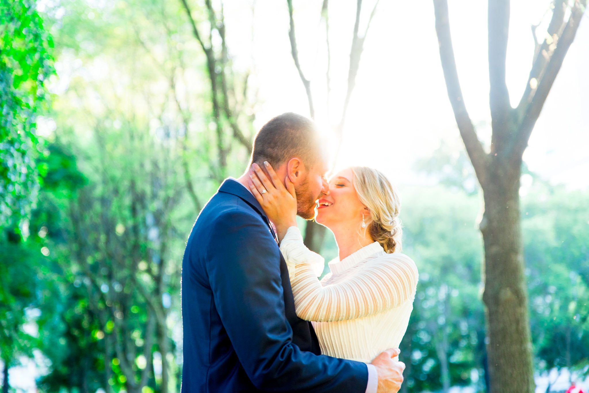 man in navy suit and woman in white dress go in for a kiss at sunset in the trees at Millennium Park