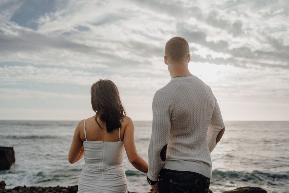 A couple stands together looking out at the ocean horizon in matching white outfits.