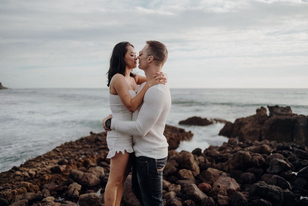 A romantic moment captured among coastal rocks with crashing waves and cloudy skies in the background.