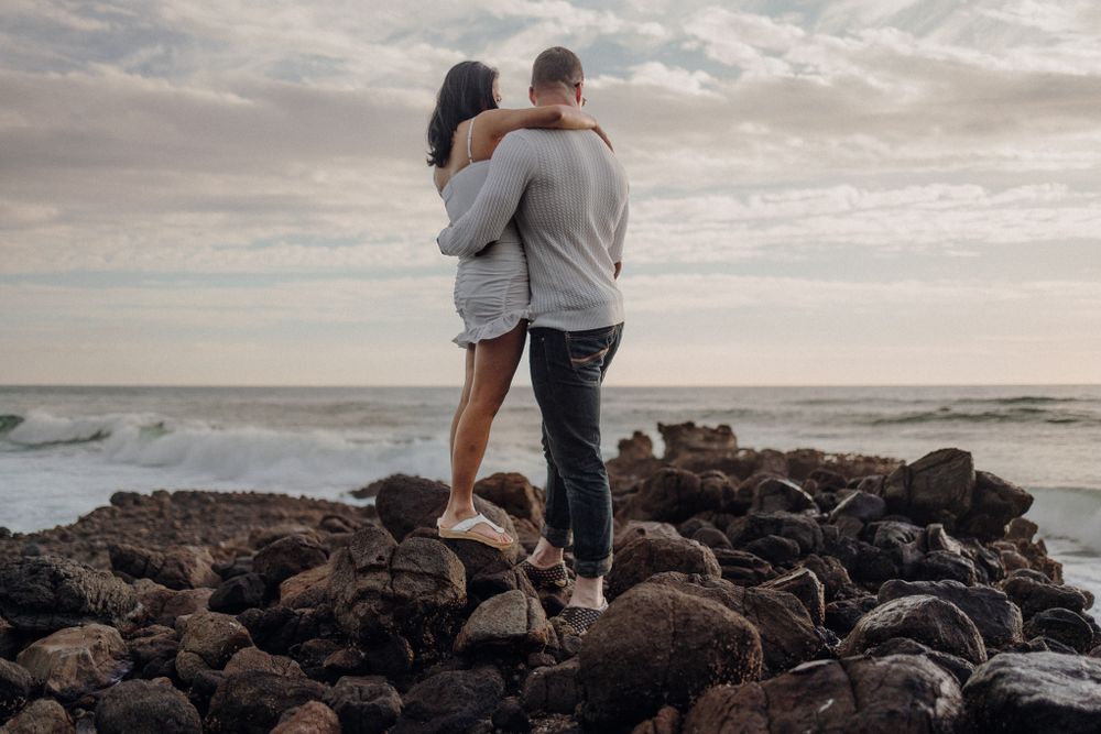 Two people stand close together on rocky coastal terrain with ocean waves in the background.