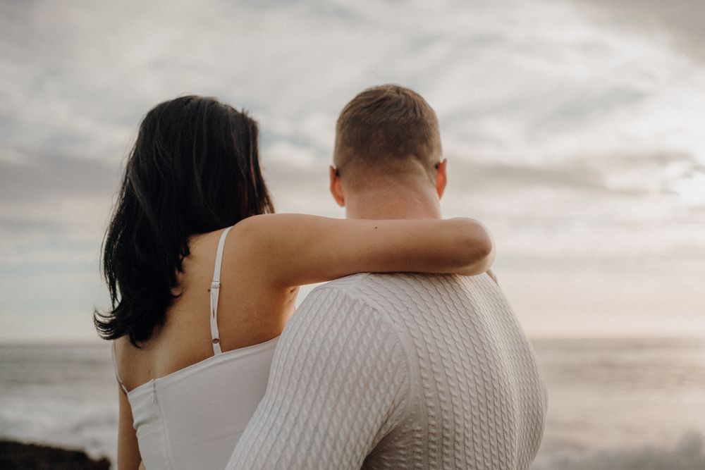 A couple in white clothing shares a tender moment overlooking the ocean at dusk.