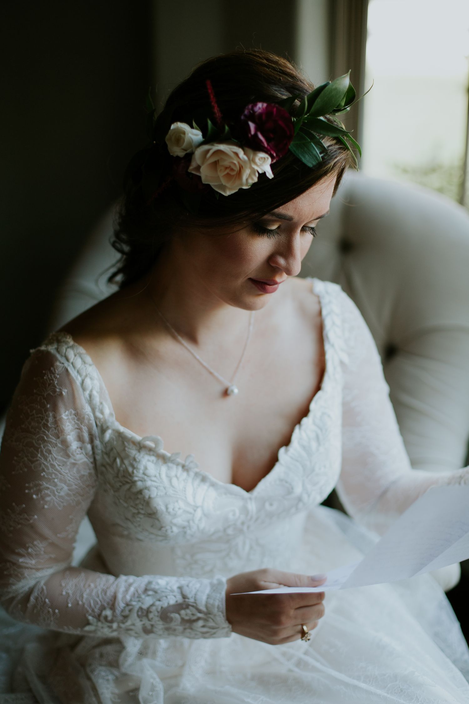 A bride wearing a floral crown and lace dress sits thoughtfully in a white chair.