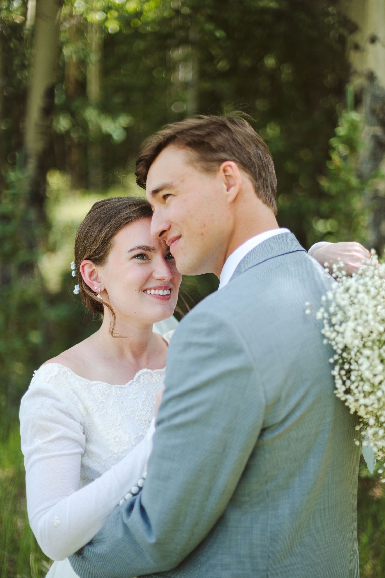 A romantic wedding portrait shows a couple sharing a tender moment amongst trees and baby's breath flowers.