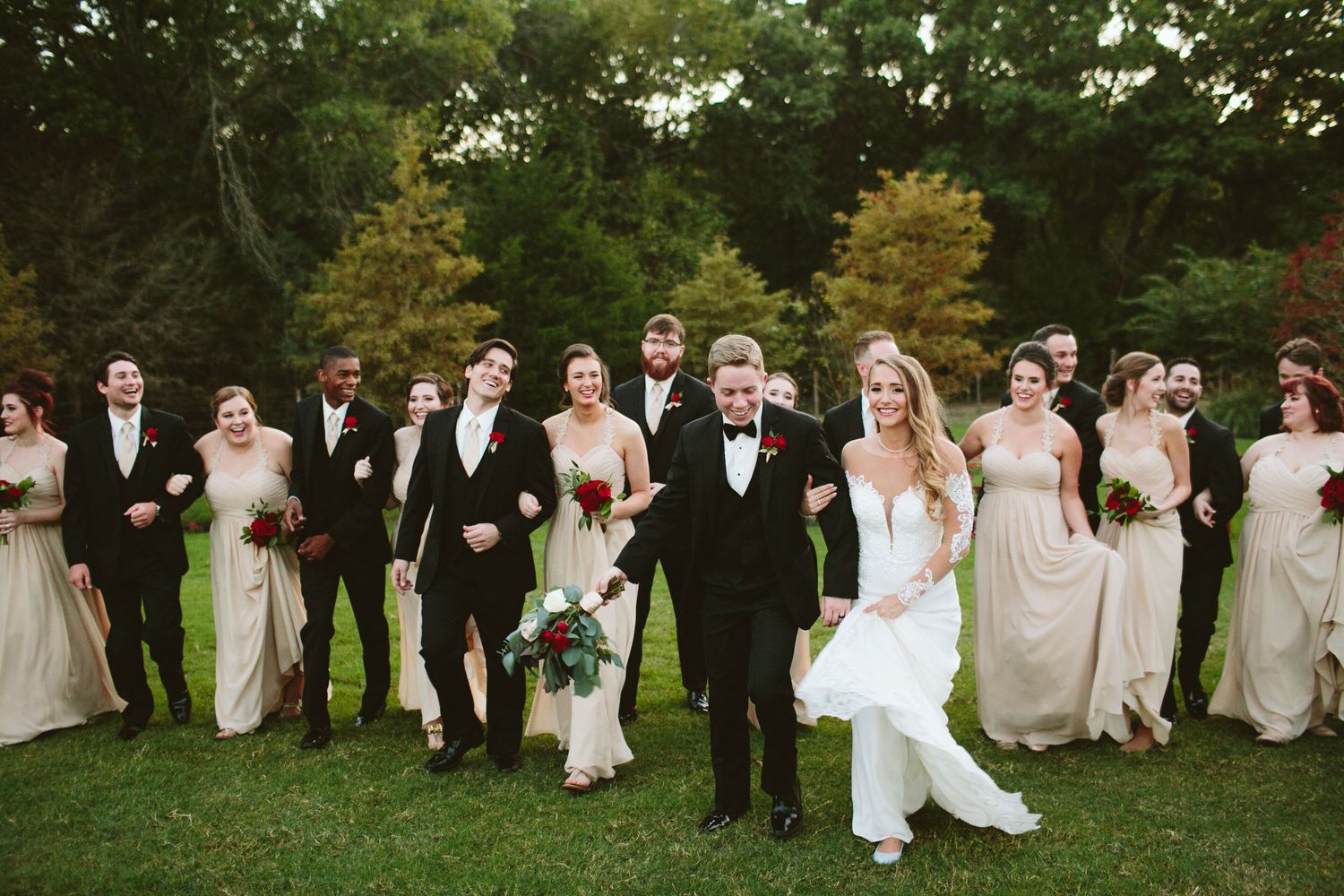 A wedding party poses outdoors on green grass with bridesmaids in neutral dresses and groomsmen in dark suits.