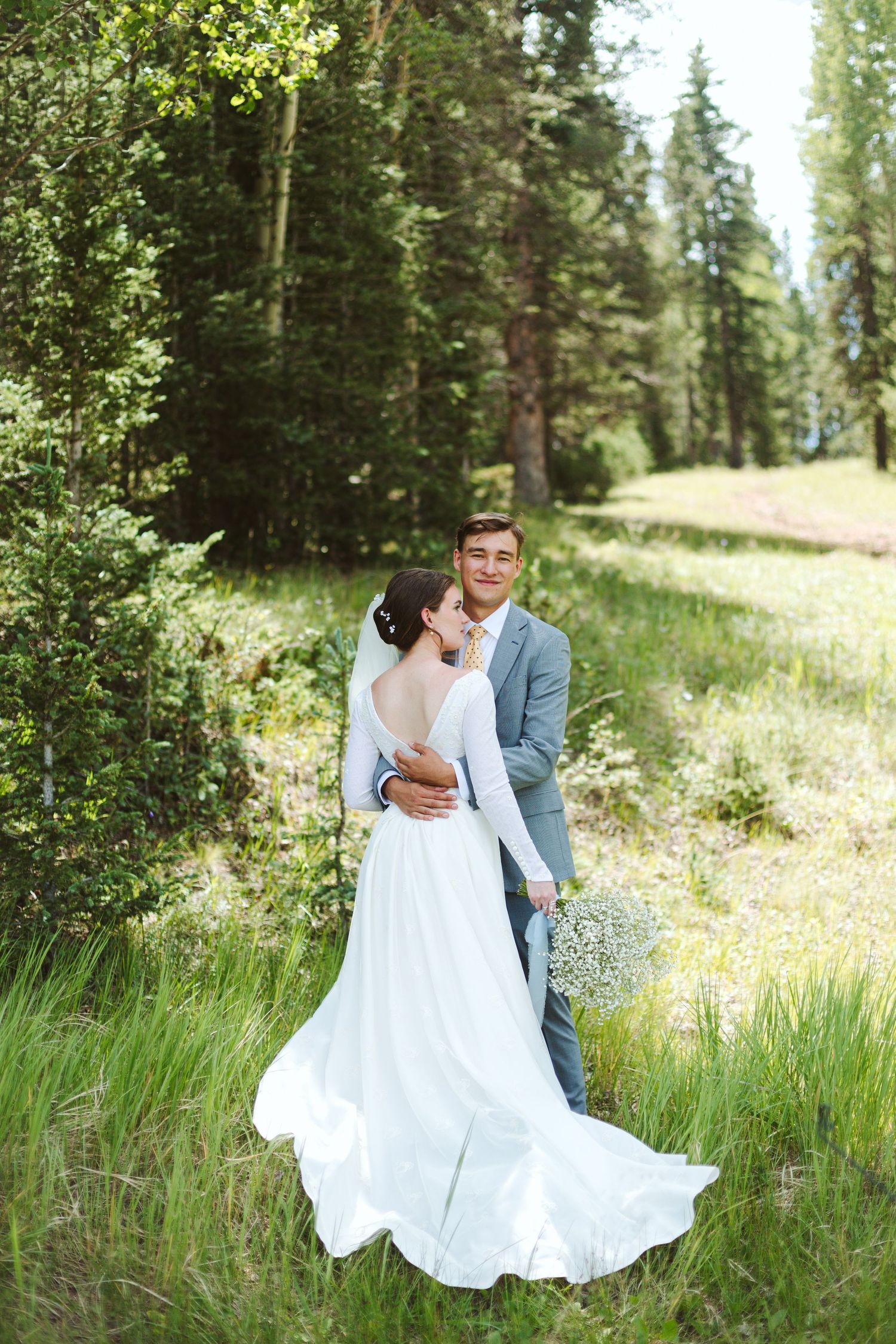 A couple shares an intimate embrace in a forest meadow with the bride wearing a flowing white gown.