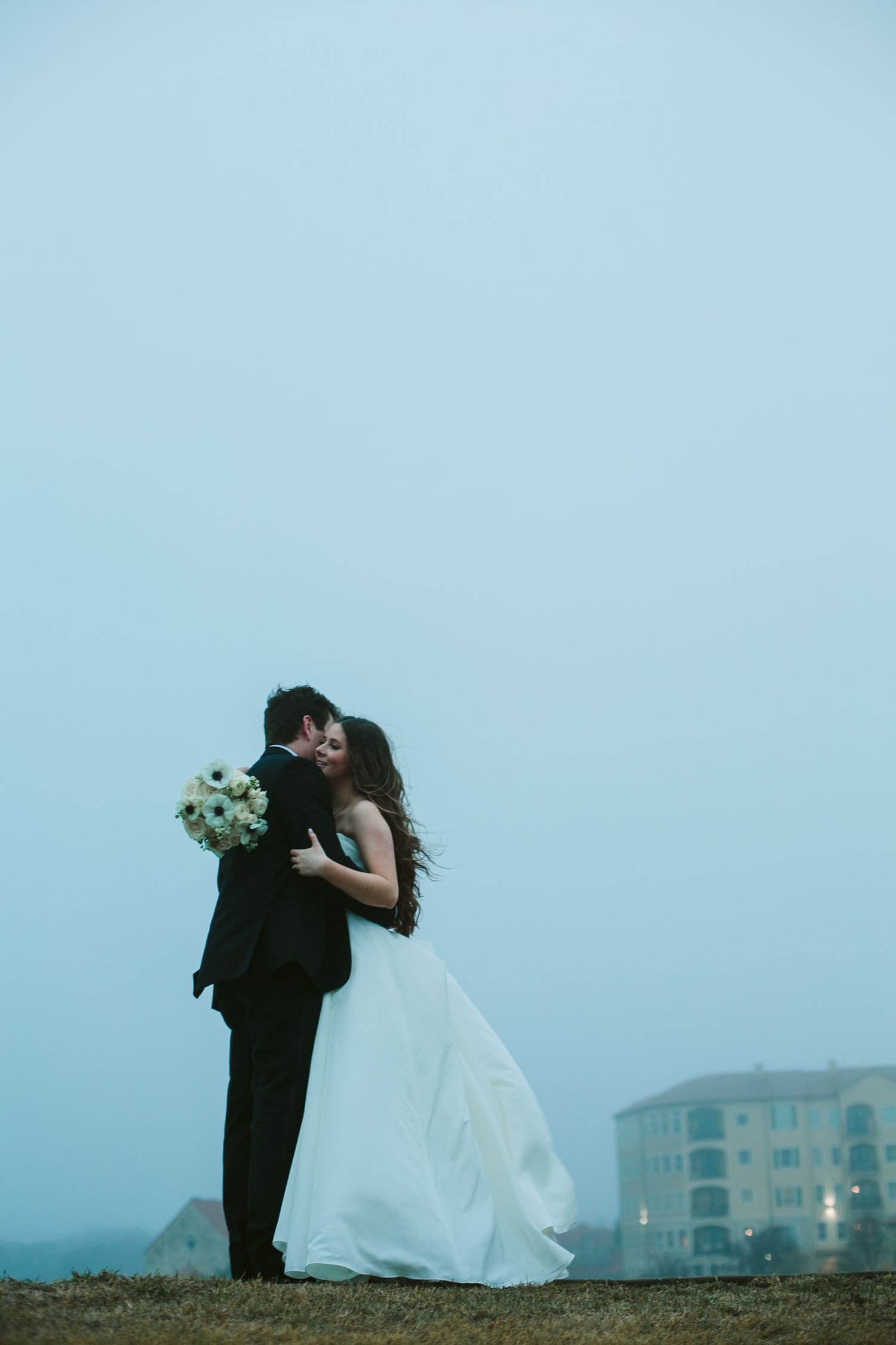 A silhouetted couple shares a romantic embrace against a misty twilight sky near coastal buildings.