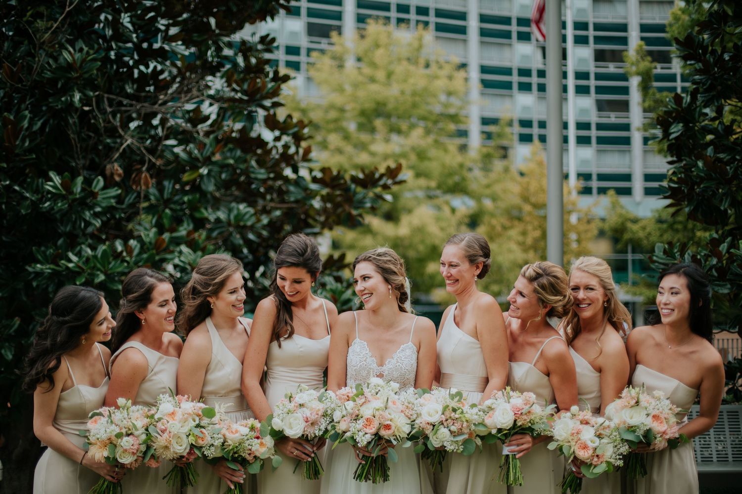 Bridesmaids in soft neutral dresses pose together holding matching floral bouquets outdoors.