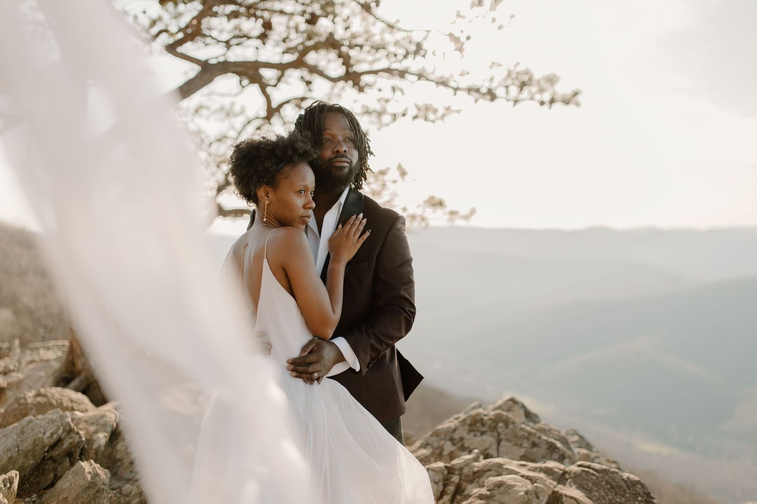 A romantic wedding couple embraces on a rocky mountain cliff at sunset.