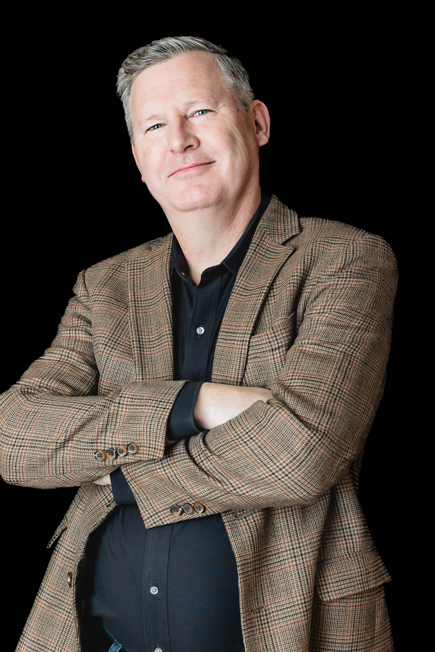Studio portrait against black background featuring person in plaid blazer with arms crossed.