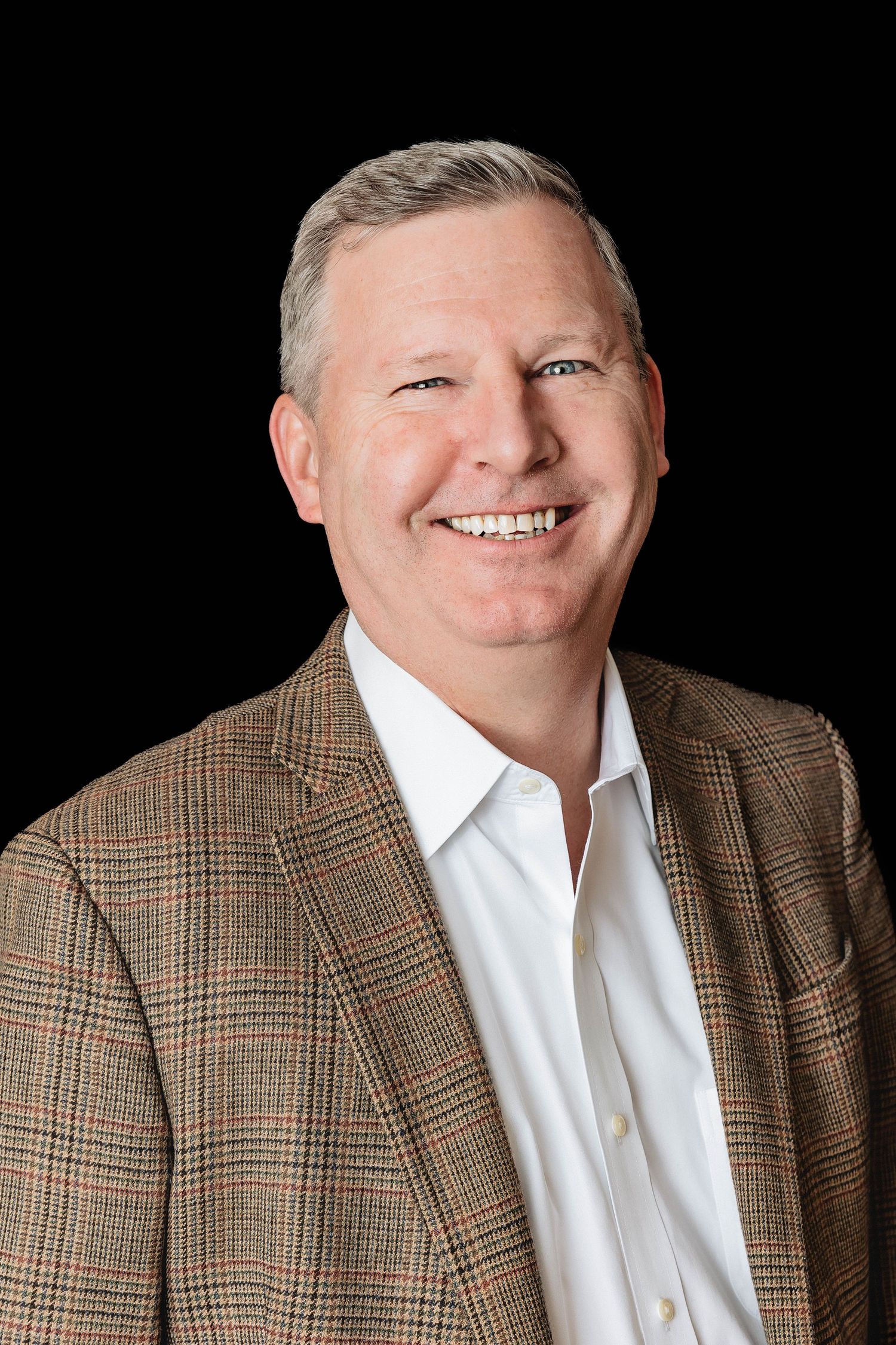 Professional headshot against black background showing person in brown plaid blazer and white shirt.