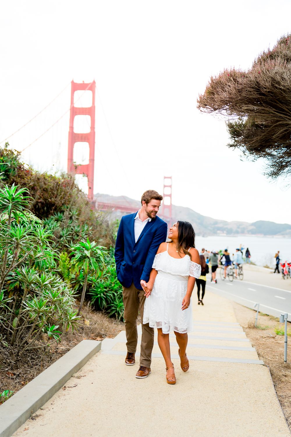 man in navy suit and woman in white dress holding hands laughing at Golden Gate Park engagement pictures