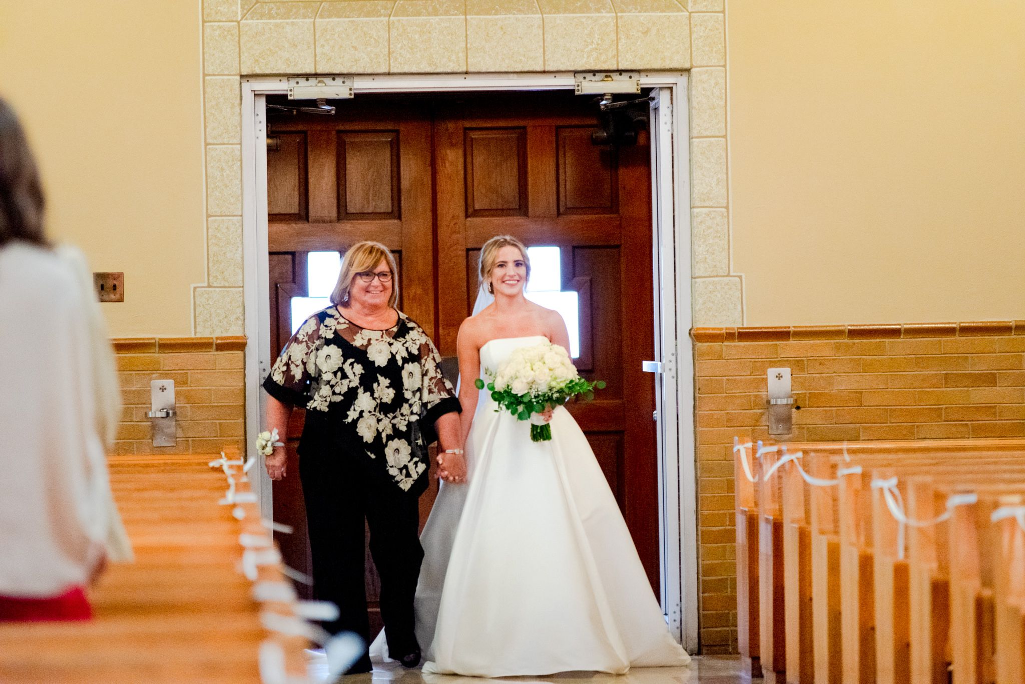 bride and mother of the bride walking down the aisle of the church both smiling and bride holding white bouquet
