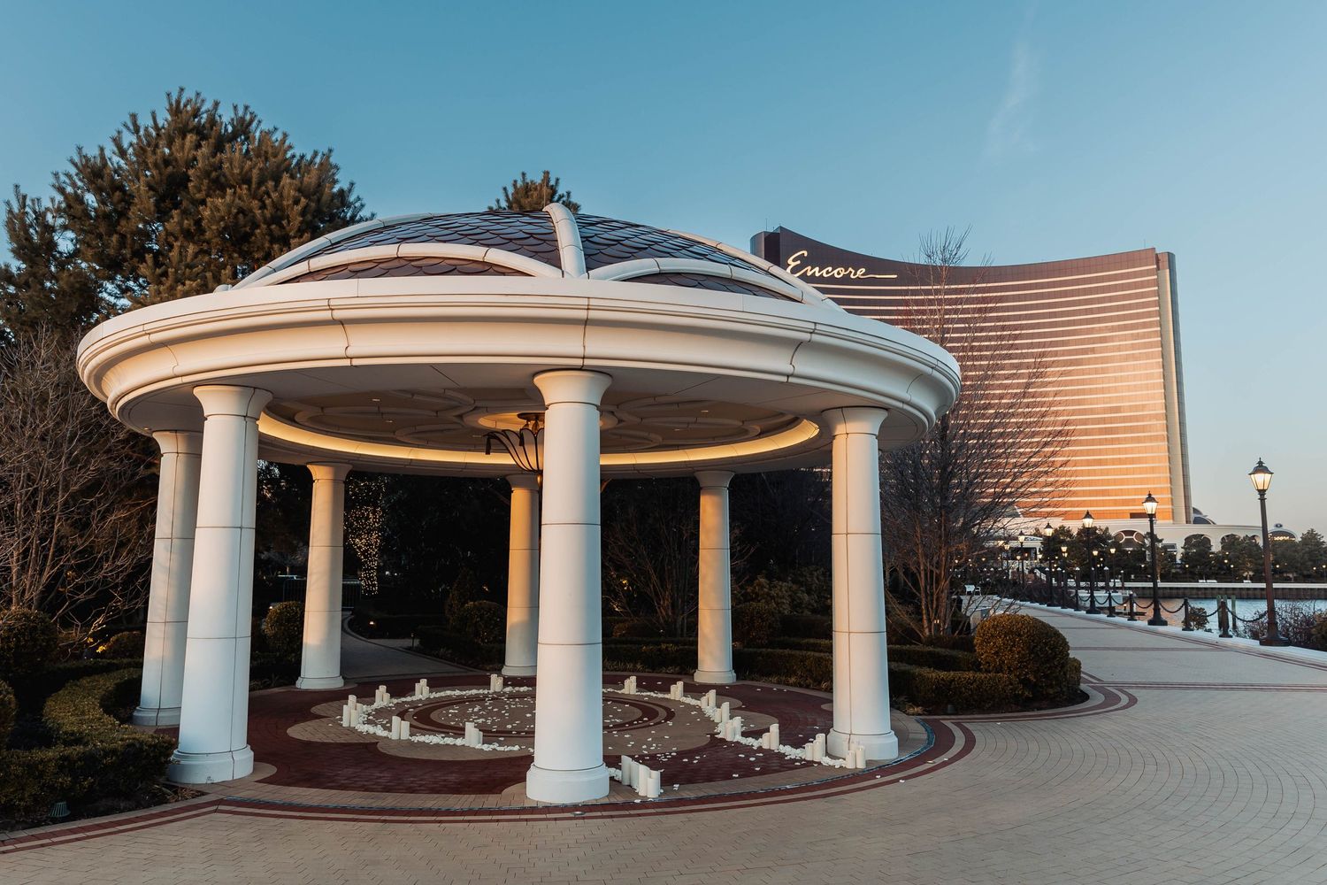 Circular gazebo with white columns lit up at dusk near modern buildings.